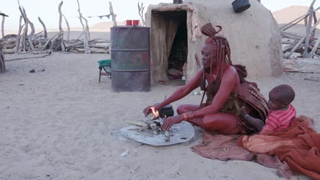 4K-view-of-Himba-woman-in-traditional-dress-with-young-child,-putting-a-small-pot-on-a-fire-outside-their-hut-within-their-small-compound,-Namibia