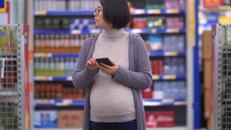 Mujer-asiática-embarazada-de-compras-en-el-mercado-mirando-al-teléfono