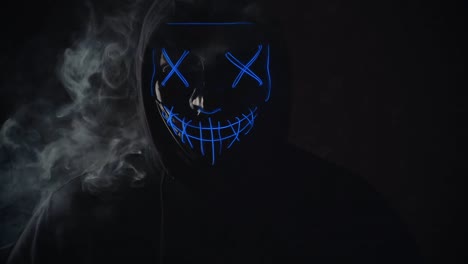 Man-with-lighting-neon-glow-mask-in-hood-and-vape-vapour-from-mask-on-black-background.-Halloween-and-horror-concept