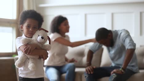 Unglücklicher-kleiner-afrikanischer-Sohn-umarmt-Spielzeug,-während-Eltern-streiten