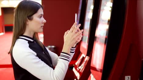 Teenager-buying-movie-ticket-from-vending-machine-at-cinema.-Woman-using-mobile,-making-photos