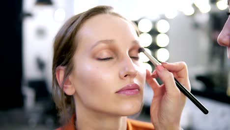 Close-up-of-a-model's-face-during-the-make-up-process.-Make-up-master-applying-light-brown-eyeshadows.-Blurred-view-of-white-lamps-on-background