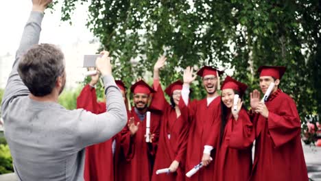 Stolzer-Vater-ist-von-Absolventen-mit-Smartphone-fotografieren,-während-junge-Menschen-posieren-sind,-winken-Hände-mit-Diplomen-und-Gestikulieren.-Konzept,-Technik-und-Bildung.