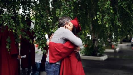 Padre-amoroso-es-felicitar-a-su-hija-en-el-día-de-la-graduación,-personas-abrazándose-y-riendo-al-aire-libre-mientras-que-otros-graduados-son-visibles-en-el-fondo.