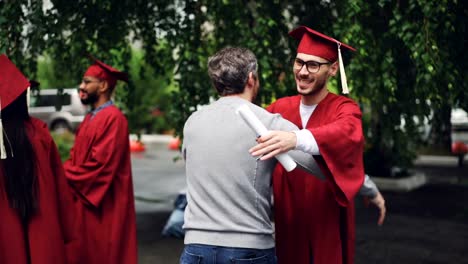 Smiling-graduating-student-is-shaking-his-father's-hand-and-hugging-him,-young-man-in-glasses-is-wearing-hat-and-gown-and-holding-diploma.-Education-and-success-concept.