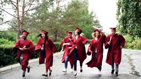 Slow-motion-of-emotional-graduates-happy-girls-and-guys-running-with-diplomas-and-laughing,-pretty-girl-is-taking-off-mortar-board-and-waving-it.-Small-rain-is-visible