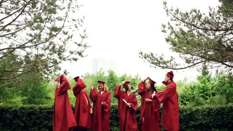 Slow-motion-of-happy-graduates-taking-off-mortarboard-caps,-throwing-in-the-air,-laughing-and-catching-them-then-jumping.-Education,-emotions-and-modern-youth-concept.