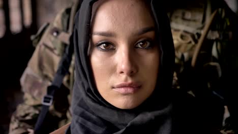 Close-portrait-of-young-muslim-woman-in-hijab-looking-at-camera,-armed-soldier-with-gun-standing-behind-woman,-military-concept