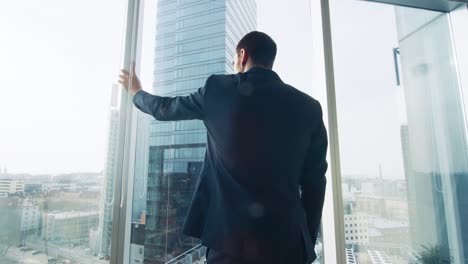 Thoughtful-Businessman-Wearing-Suit-Standing-in-His-Office,-Looking-out-of-the-Window-and-Contemplating-Next-Big-Business-Contract.-Major-City-Business-District-with-Panoramic-Window-View.-Blue-Colors.