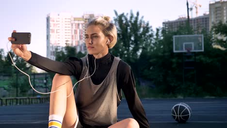 Beautiful-blonde-female-basketball-player-taking-selfie-and-sitting-on-ground,-men-playing-in-background,-court-in-open-air