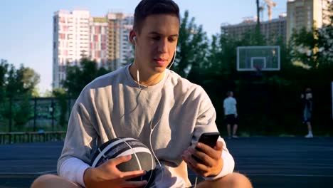 Joven-guapo-sentado-en-la-cancha-de-básquet-y-tecleando-en-el-teléfono,-mirando-a-cámara,-sosteniendo-la-bola,-la-gente-tocando-fondo