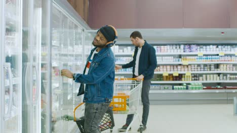 En-el-supermercado:-elegante-chico-afroamericano-con-cesta-de-la-compra-elige-los-productos-en-la-sección-de-congelados-de-la-tienda.-Abre-refrigerador-puerta-de-cristal-grande.-Cámara-lenta.