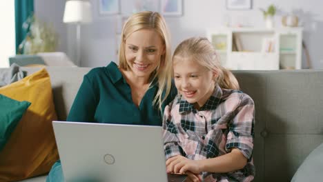 Beautiful-Young-Mom-and-Her-Cute-Little-Daugther-Use-Laptop-while-Sitting-on-a-Sofa-at-Home.-Family-Spending-Time-Together-Watching-Videos-and-Cartoons-on-Computer.