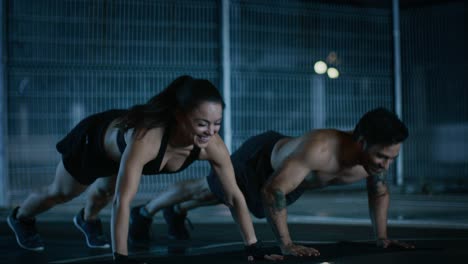 Smiling-Happy-Athletic-Fitness-Couple-Doing-Push-Up-Exercises-and-Giving-High-Five.-Workout-is-Done-in-a-Fenced-Outdoor-Basketball-Court.-Night-Footage-After-Rain-in-a-Residential-Neighborhood-Area.