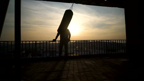 Silhouette-of-boxing-bag-while-swinging,-sunset,-man-comes-and-looks-over-fence,-sportsman-practicing,-power-training,-strong-guy-hard-exercising,-strength-exercises,-workout,-handheld,-sunny-day.