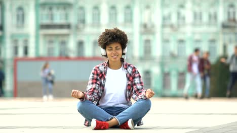 Smiling-woman-listening-to-music-and-sitting-in-lotus-position-on-urban-street