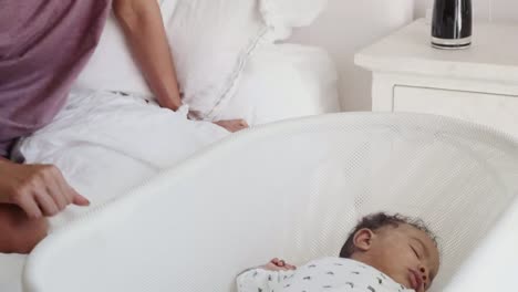 Madre-Millennial-sentada-en-la-cama-mirando-a-su-bebé-recién-nacido-durmiendo-en-una-cuna,-tiro-inclinado
