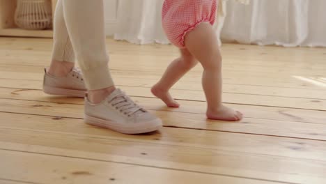 Tracking-close-up-shot-of-unrecognizable-caring-mother-supporting-her-barefoot-baby-girl-trying-to-walk-on-hardwood-floor