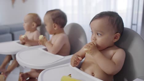 Gemischte-Rasse-Baby-Triplets-sitzen-in-Hochstühlen-und-Essen-Bananen