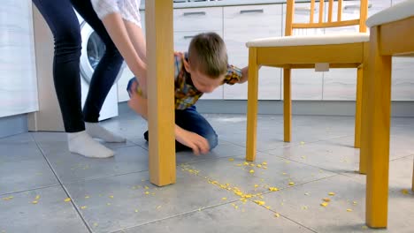 Mama-schimpft-ihren-Sohn-für-verstreutes-Essen-auf-dem-Küchenboden-und-lässt-ihn-aufräumen.-Maisflocken-gemeinsam-vom-Boden-säubern.