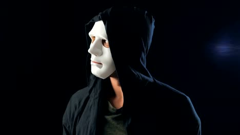 Anonymous-man-in-a-white-mask-covering-his-face-and-a-black-hood-turns-and-looks-at-the-camera.-Black-dark-background.-Concept-of-a-member-of-a-secret-organization