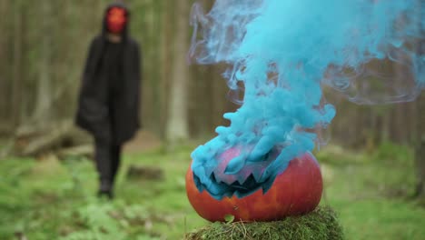 Person-goes-in-mask-and-coak-in-the-forest-on-the-halloween-with-smoky-pumpkin