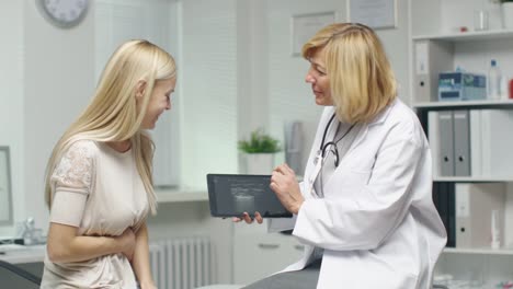 Mid-Adult-Female-Doctor-Consults-Young-Female-by-Showing-to-Her-Tablet-Computer-with-X-Ray.-Frau-hodelt-ihren-Stomach.-Beide-Lächeln