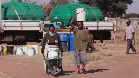 Zwei-afrikanische-Frau-Wasser-aus-Tankfahrzeugen-in-Kunststoffeimern-zu-sammeln-und-zu-Fuß-zurück-zu-ihren-Häusern