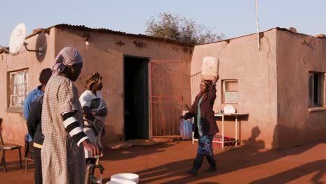 Mujer-africana-al-llegar-a-una-casa-para-recoger-agua-en-un-balde-plástico,-mientras-que-otros-están-parados-en-línea-y-esperan-su-turno
