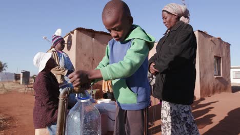 Joven-africano-que-recoge-agua-de-un-grifo-mientras-mujer-en-fila-para-recoger-agua-en-envases-de-plástico-debido-a-la-sequía-en-África-del-sur