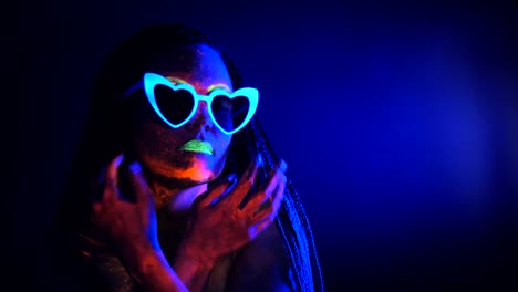 Fashion-sexy-dancer-with-heart-shaped-glasses-in-neon-light.-Fluorescent-makeup-glowing-under-ultraviolet-light.-Night-club,-party,-psychedelic-concepts.-Mysterious-woman-with-UV-painting