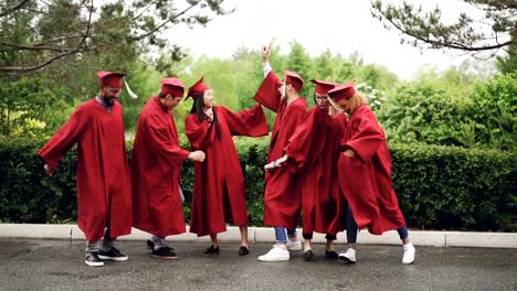 Cámara-lenta-de-juguetonas-chicas-y-chicos-graduar-estudiantes-bailando,-riendo-y-divirtiéndose-en-el-día-de-la-graduación.-Día-hermoso-de-verano,-árboles-verdes-son-accesibles.