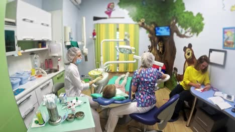 Child-with-a-mother-at-a-dentist's-reception.-The-girl-lies-in-the-chair,-behind-her-mother.-The-doctor-works-with-an-assistant.-Procedure-for-drilling-a-tooth.-Setting-a-temporary-seal.