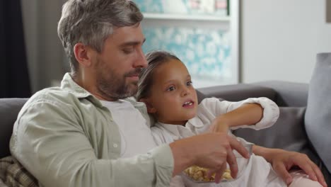 Papa-und-kleines-Mädchen-vor-dem-Fernseher-auf-Sofa-und-essen-Popcorn