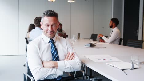 Portrait-Of-Mature-Businessman-Sitting-In-Modern-Boardroom-With-Colleagues-Meeting-Around-Table-In-Background-Shot-In-Slow-Motion