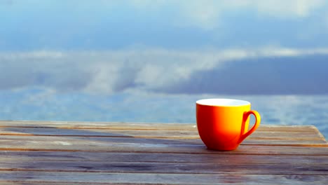 Orange-cup-of-coffee-on-brown-wooden-table-on-beach-with-surf-of-blue-sea-in-background.
