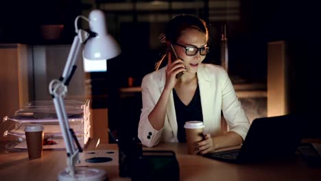 Cute-girl-company-manager-is-talking-on-mobile-phone-and-using-laptop-sitting-at-desk-in-dark-office-and-smiling-enjoying-her-job-and-holding-to-go-coffee.