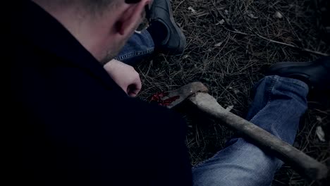 Male-psychopath-sits-and-picks-up-dried-blood-on-axe-in-woods