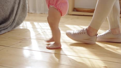 Tracking-close-up-shot-of-unrecognizable-caring-mother-helping-her-barefoot-baby-daughter-walking-towards-sofa-at-home