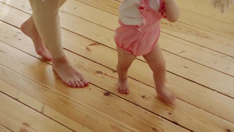 Adorable-baby-girl-learning-to-walk-on-hardwood-floor-at-home-supported-by-her-loving-mother,-tracking-shot