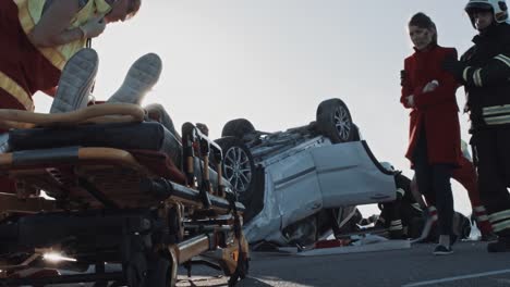 Car-Crash-Traffic-Accident:-Firefighter-Leads-Mother-to-a-Child-Lying-on-Stretchers-Whom-Paramedics-Trying-to-Save-/-Resuscitate.-Rollover-Vehicle-with-Other-Trapped-Passengers-in-the-Background