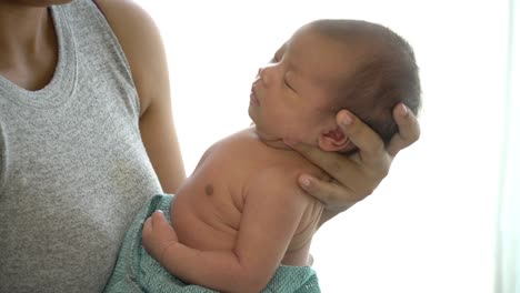 4K-Medium-close-up-shot-of-young-adult-asian-woman-mother-standing-near-window-in-home-bedroom-and-carrying-her-sleeping-nap-newborn-baby-girl-daughter.-Mother-day-and-baby-health-care-concept.