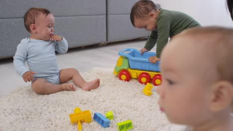 Tres-niños-pequeños-jugando-en-la-alfombra-en-casa-y-la-madre-mirando