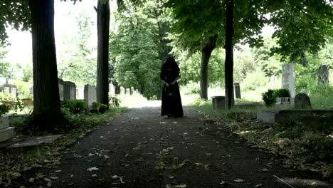 Woman-in-mourning-black-clothes-walking-slowly-on-alley-in-cemetery-holding-a-flower-crown-in-her-hand-portrait-of-sorrow-and-loneliness