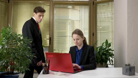 A-young-guy-in-a-suit-flirts-with-a-girl-in-the-office,-gives-her-a-green-abloko,-a-girl-works-on-a-computer,-a-red-laptop.-60-fps
