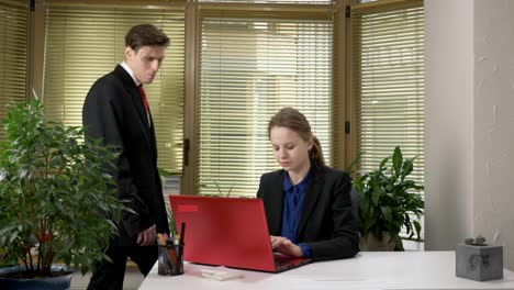 Ein-junger-Mann-in-einem-Anzug-flirtet-mit-einem-Mädchen-im-Büro,-gibt-ihr-einen-grünen-Abloko,-ein-Mädchen-auf-einem-Computer,-einem-roten-Laptop-funktioniert.-60fps