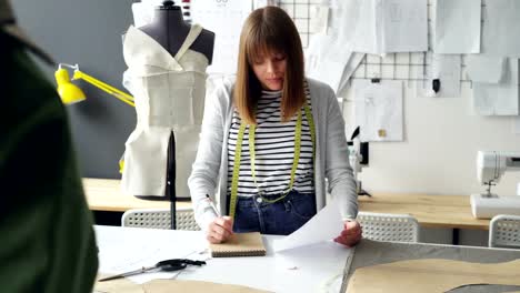 Professionelle-Schneiderin-macht-Notizen-im-Editor-und-während-der-Arbeit-am-Studio-Tisch-mit-Blick-auf-Kleidung-Skizze.-Viele-Zeichnungen,-Dummy-und-Nähmaschine-im-Hintergrund.