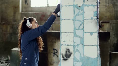 Female-graffiti-painter-is-creating-abstract-image-on-pillar-in-old-damaged-warehouse-and-listening-to-music-with-headphones.-Modern-art-and-creative-people-concept.