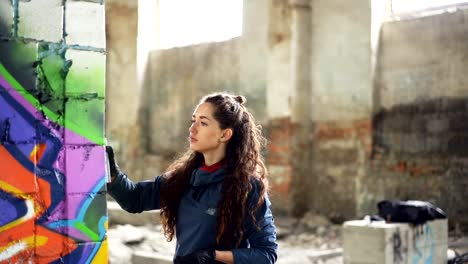 Slow-motion-of-graffiti-artist-painting-on-wall-in-abandoned-building-using-aerosol-spray-paint.-Attractive-girl-with-curly-hair-is-busy-with-her-work,-she-is-looking-at-painting.