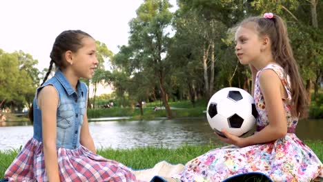 Zwei-kleine-Mädchen-im-Park-auf-dem-Rasen-einander-gegenüber-sitzen,-halten-einen-Fußball-und-aktiv-etwas-zu-diskutieren.